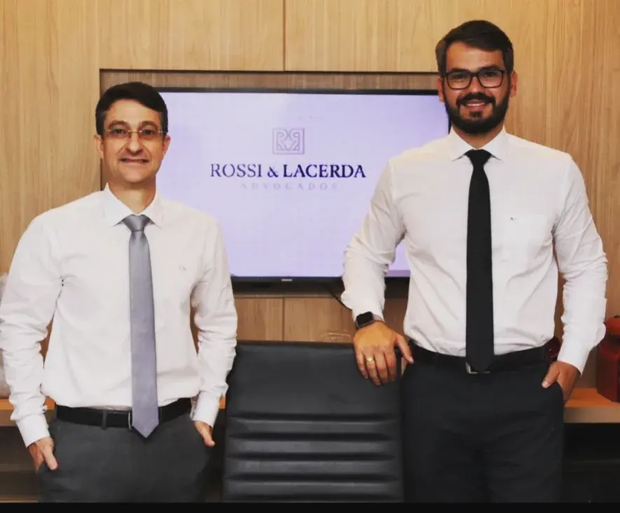 Cristiano Rossi e Deangelis Lacerda em pé na sala de reunião do escritório, diante da tela com o logo Rossi & Lacerda